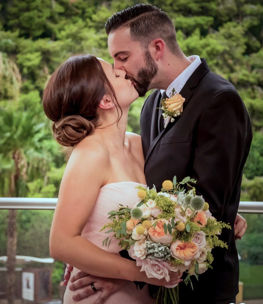 A bride and groom kissing on a balcony while holding a bouquet of flowers.
