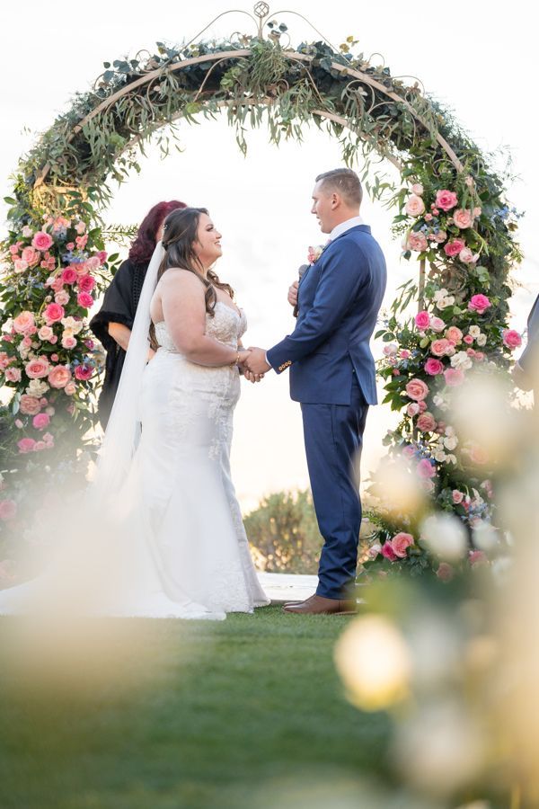 A bride and groom are holding hands during their wedding ceremony under a floral arch.
