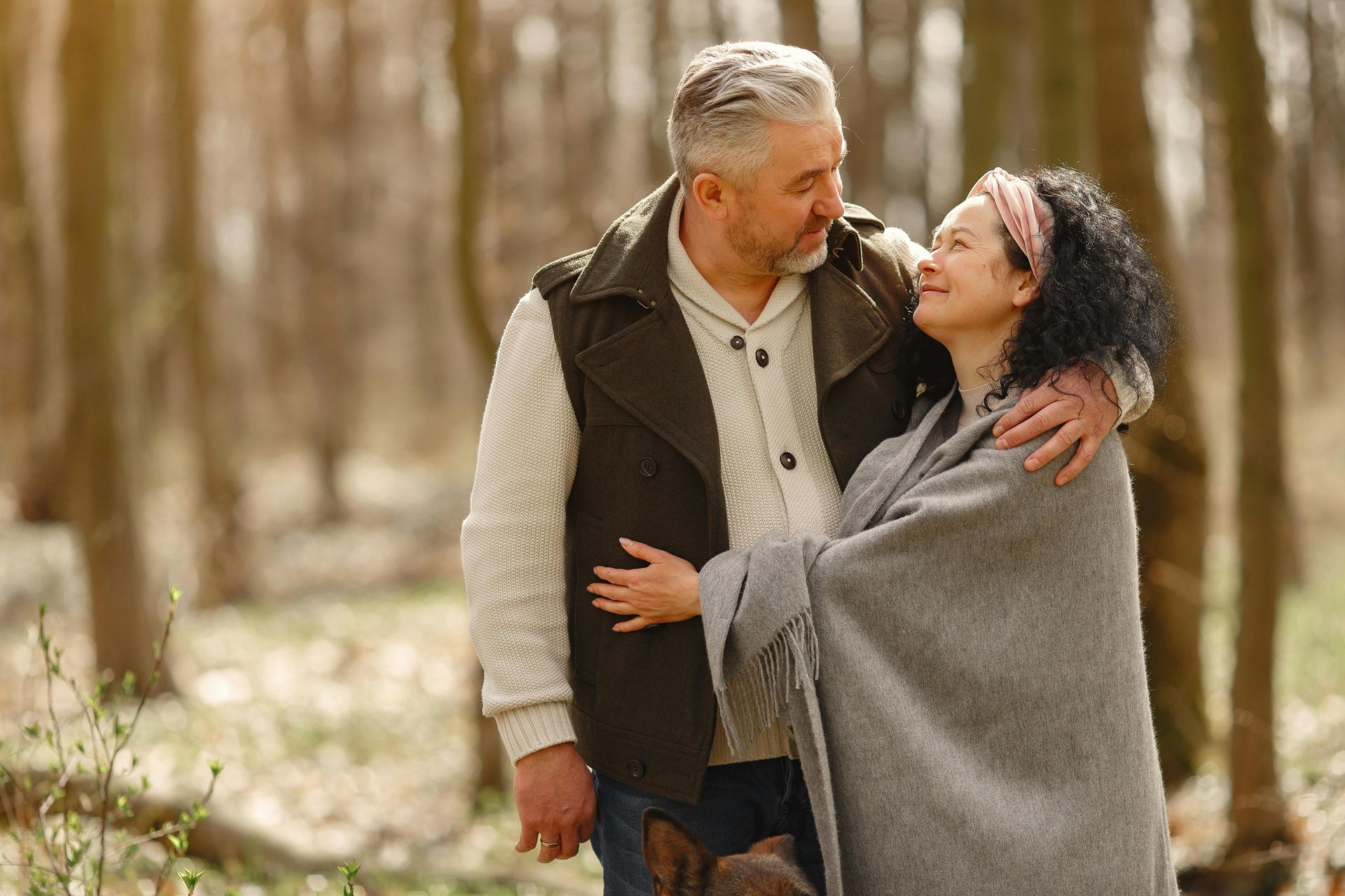 Mature couple embraces in a forest, woman wrapped in a gray blanket, man's arm around her.