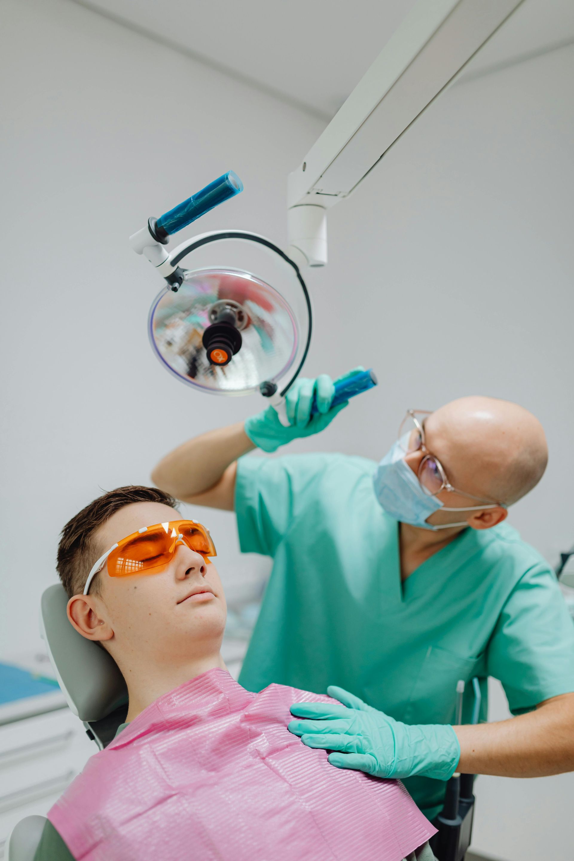 Dentist examining patient's teeth under a bright light. Patient wears orange glasses and bib.