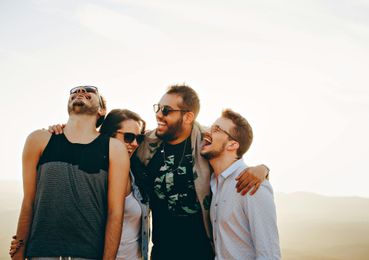 Four friends laughing with arms around each other, wearing sunglasses, in a sunny outdoor setting.