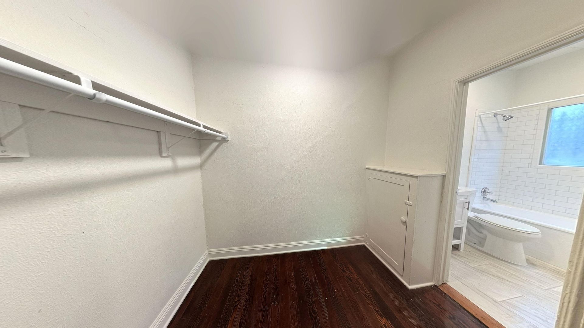 Small, empty walk-in closet with dark wood floors, white walls, and an adjacent bathroom visible through the doorway.