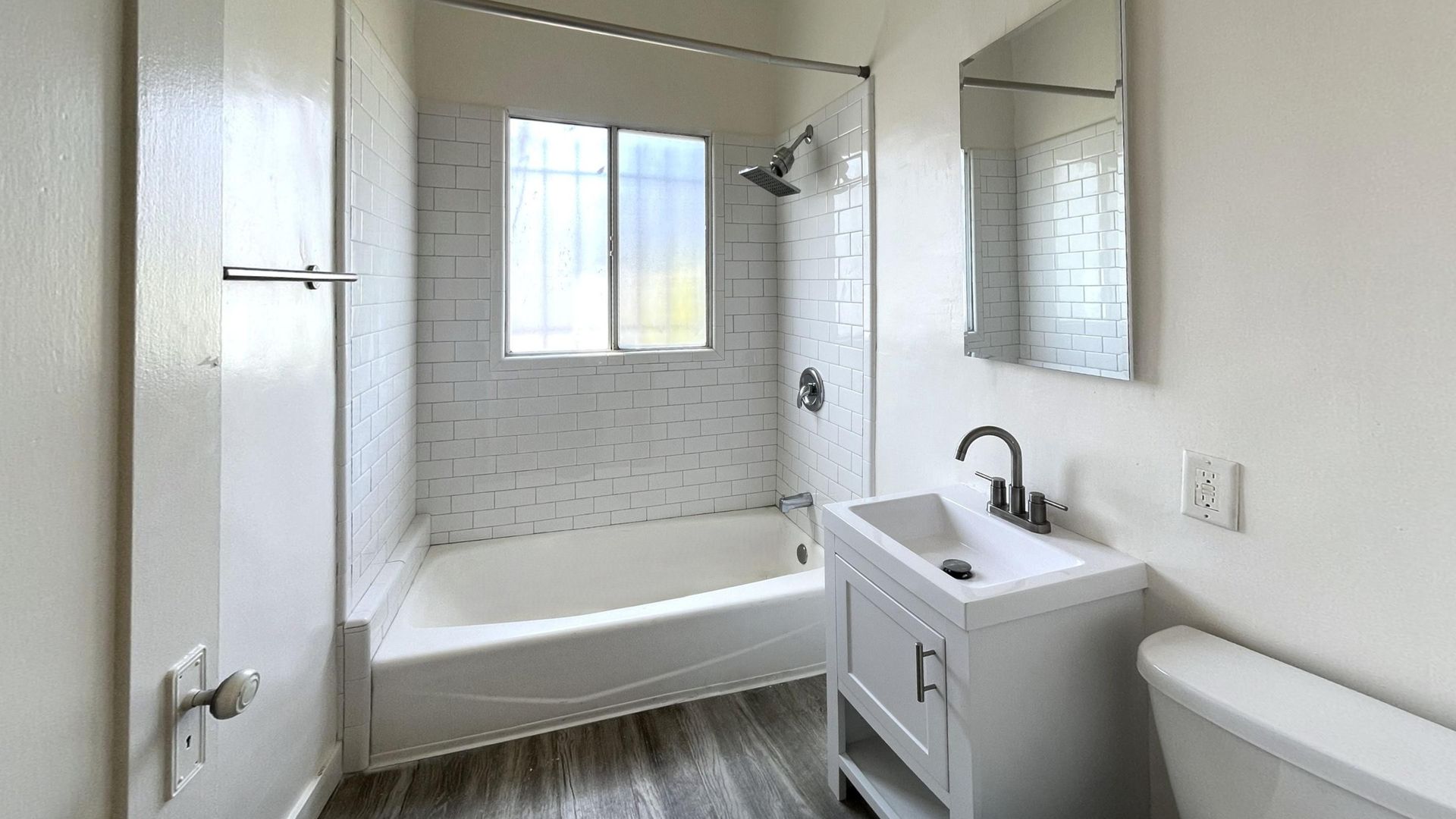 Bathroom with white walls, tub, vanity, mirror, and toilet. A window is above the tub.