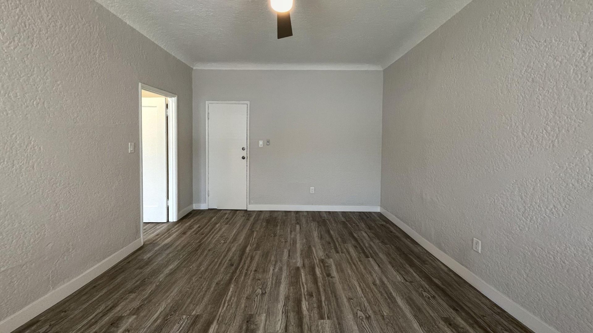 Empty room with light gray walls, dark wood-look flooring, white door, and a ceiling fan.