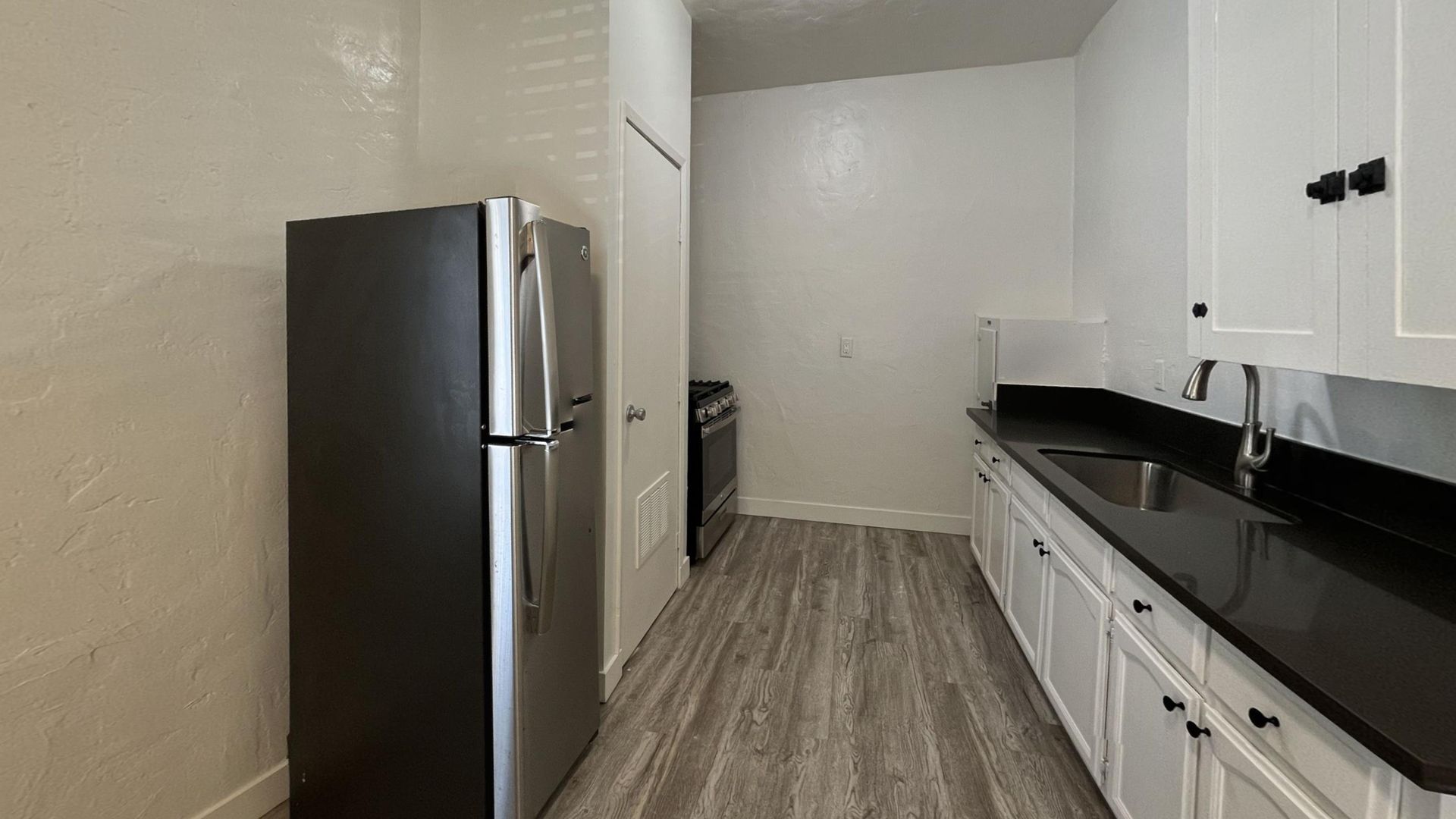 Kitchen with stainless steel refrigerator, white cabinets, black countertop, and gray wood-look flooring.