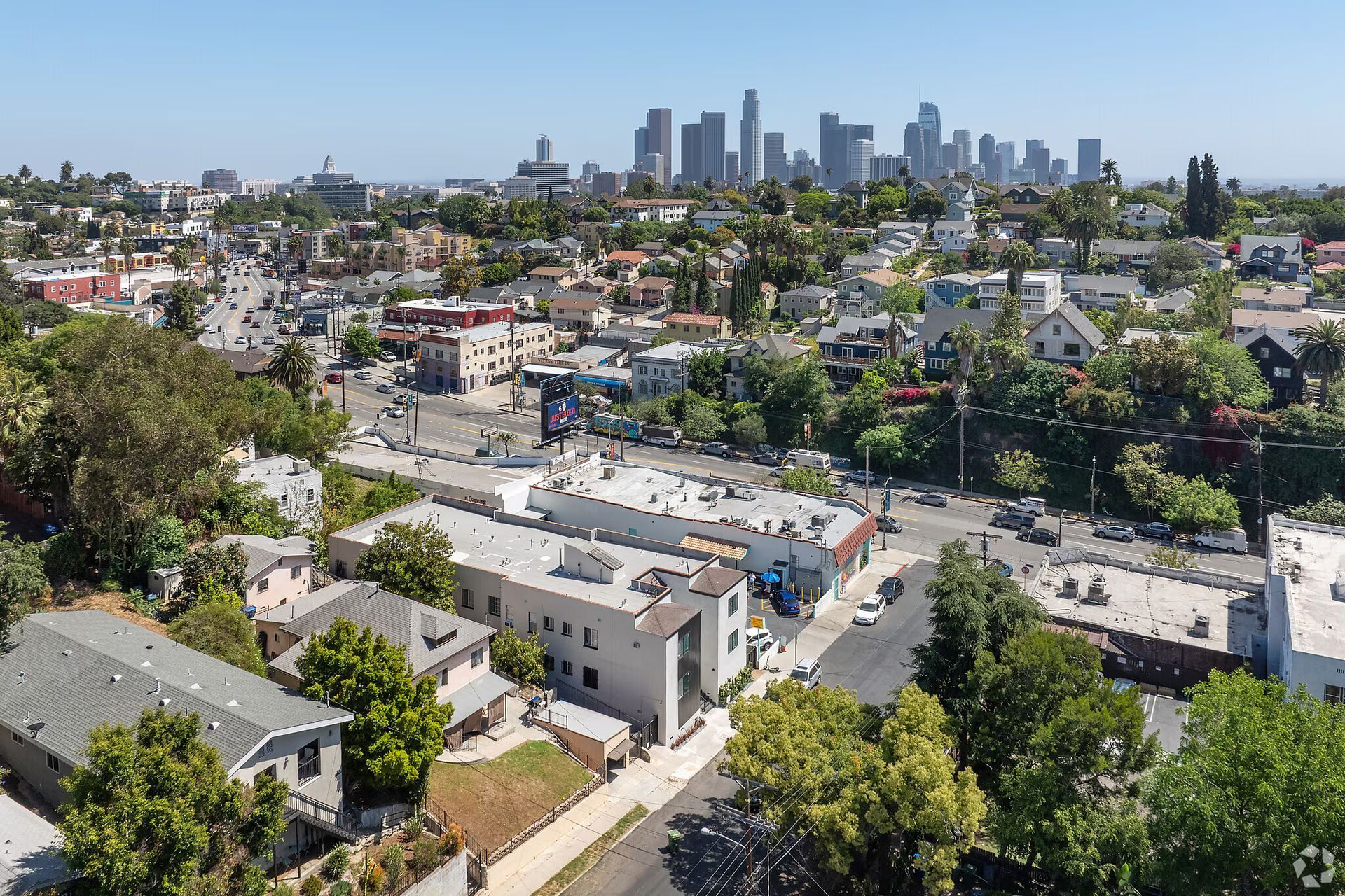 Aerial view of a Los Angeles neighborhood with downtown skyline in the background, under a blue sky.