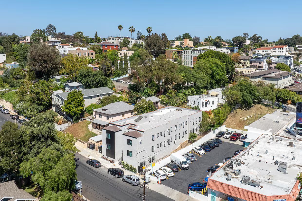 Aerial view of apartment complex with parking, nestled among other houses on a hillside, under a blue sky.