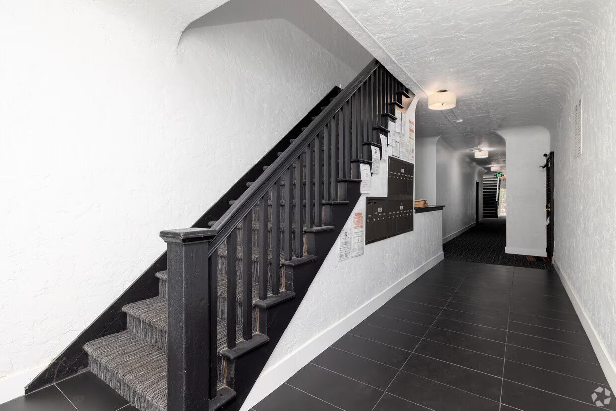 Stairwell with black railing and steps, leading up. Long hallway with black tiled floor and white walls.