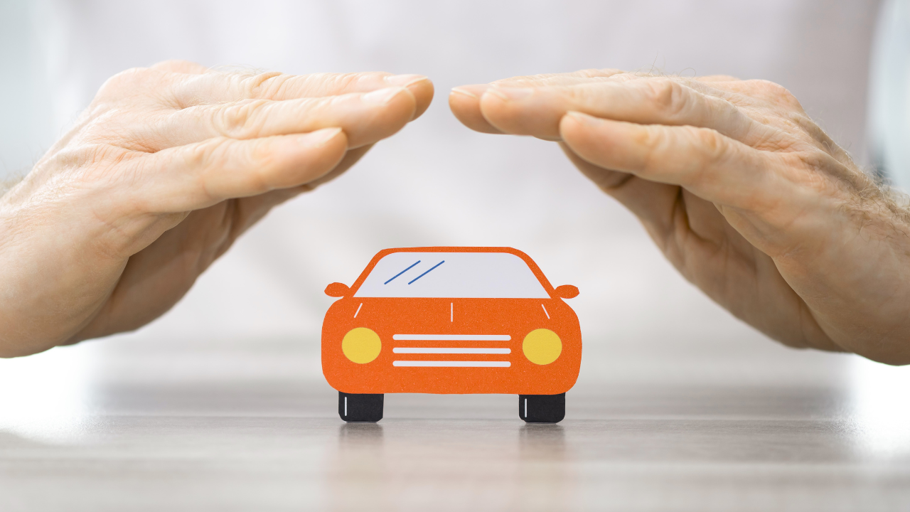 Two hands held protectively over a small, orange, paper cut-out of a car on a table.