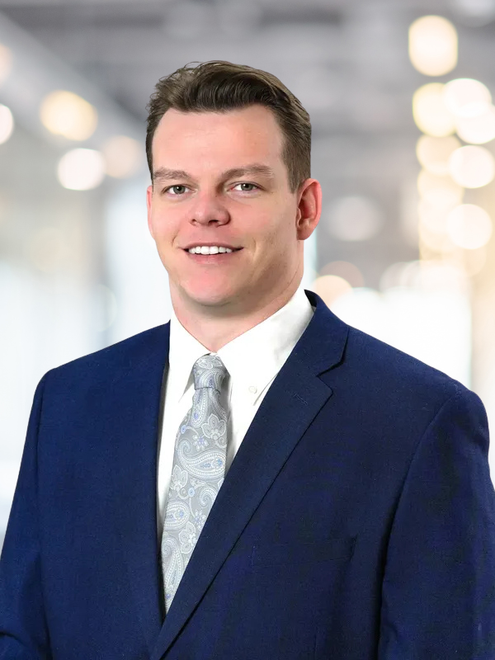 Man in a blue suit and tie smiling, blurred office background.