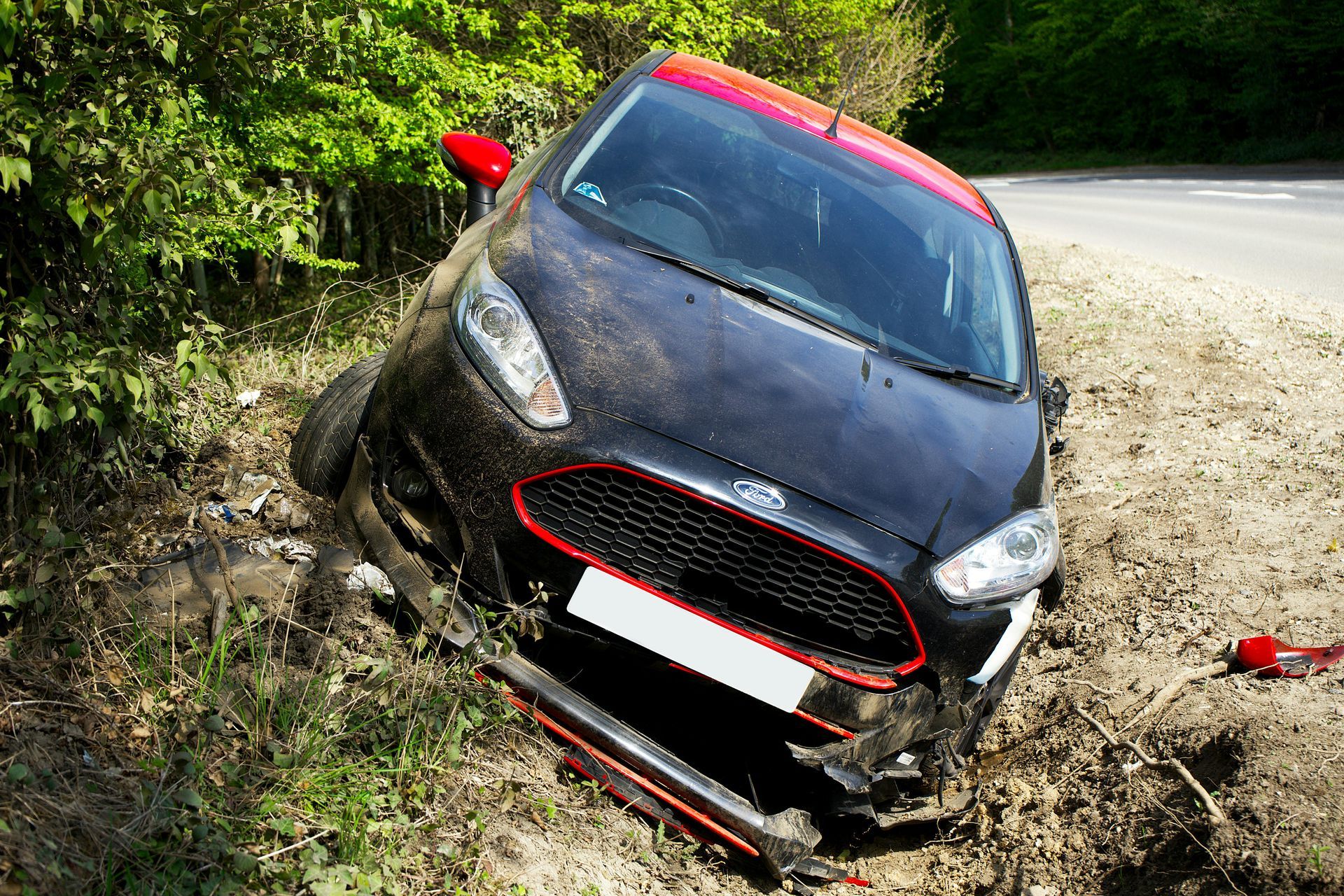 A black Ford Fiesta with red trim is crashed in a roadside ditch surrounded by trees and dirt.