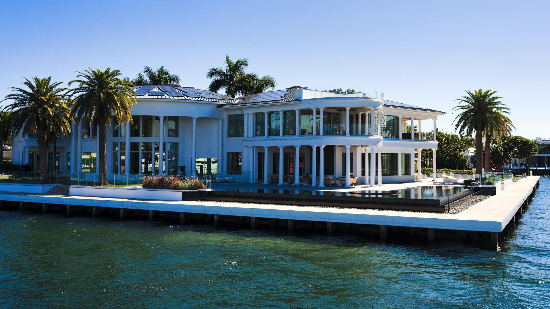 A white, two-story luxury mansion with pillars and palm trees, situated on a waterfront lot.