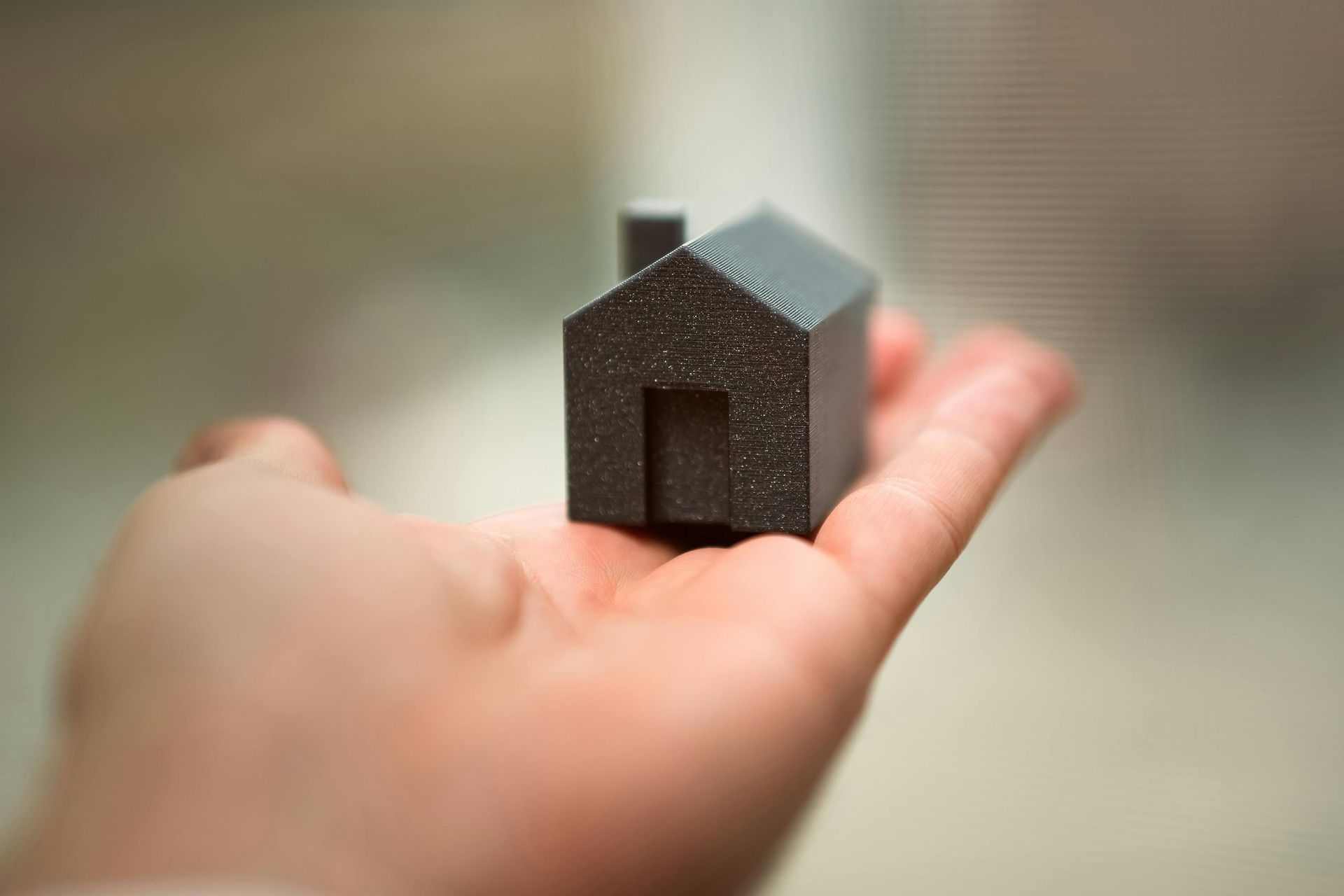Small dark miniature house resting on an outstretched hand against a blurred background