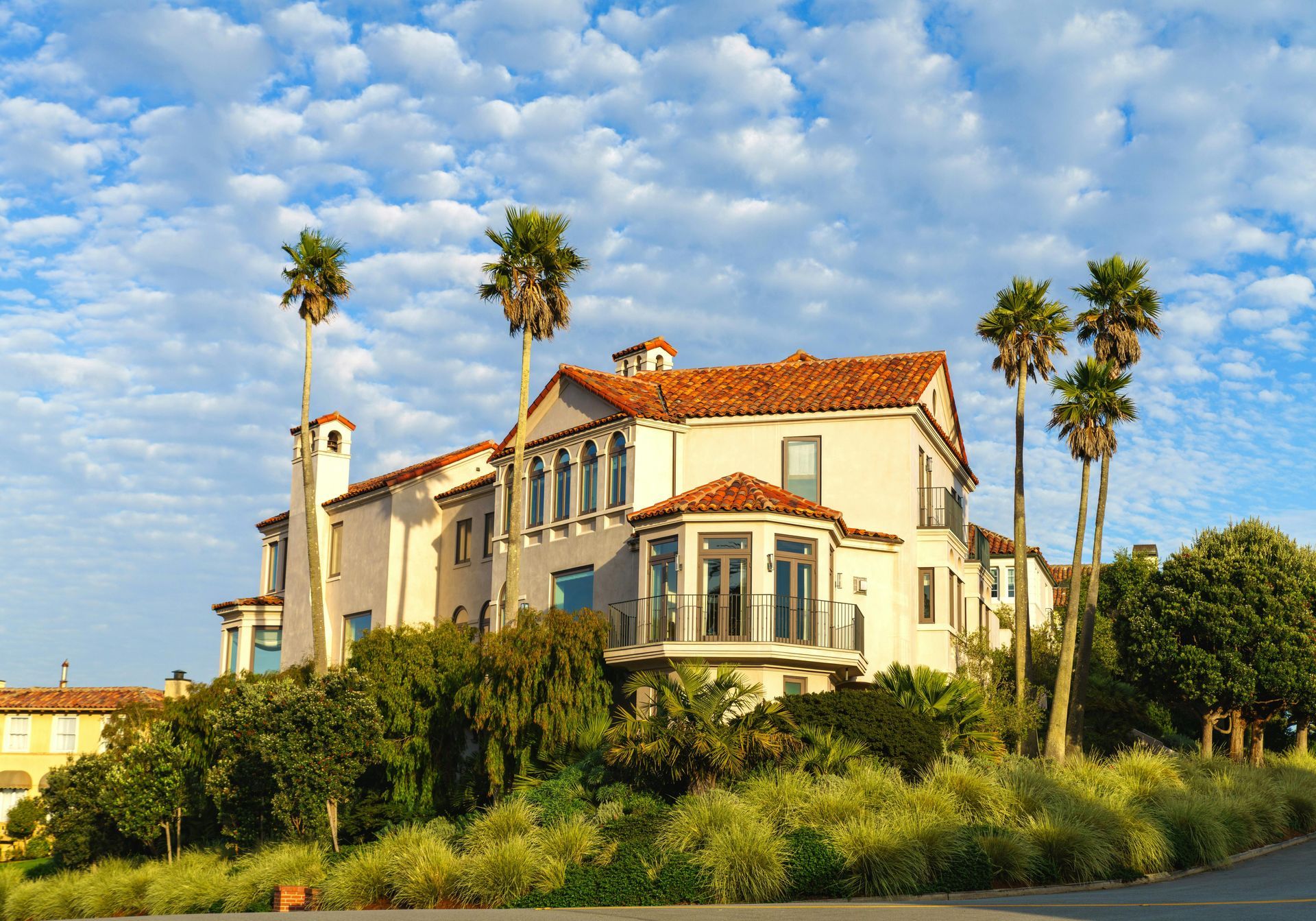 A grand, multi-story Spanish-style house with a terracotta roof, set against a bright, cloudy sky with palm trees.