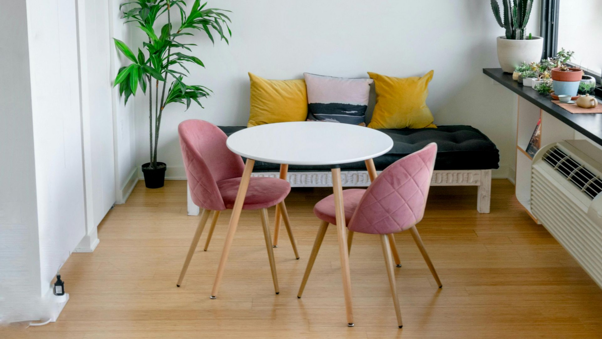A dining nook featuring a small white table with two pink velvet chairs, set against a cushioned bench with yellow pillows.