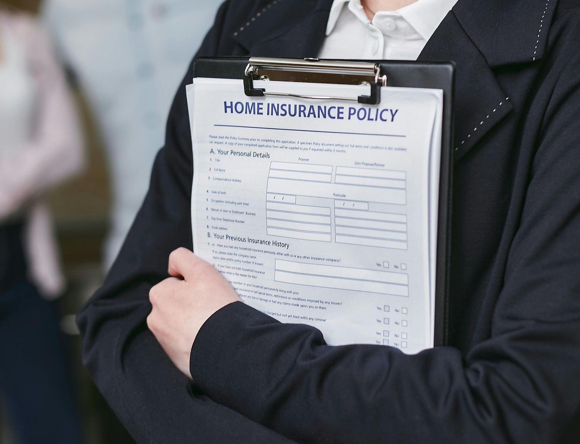 A person in a dark suit holding a clipboard with a Home Insurance Policy form against their chest.