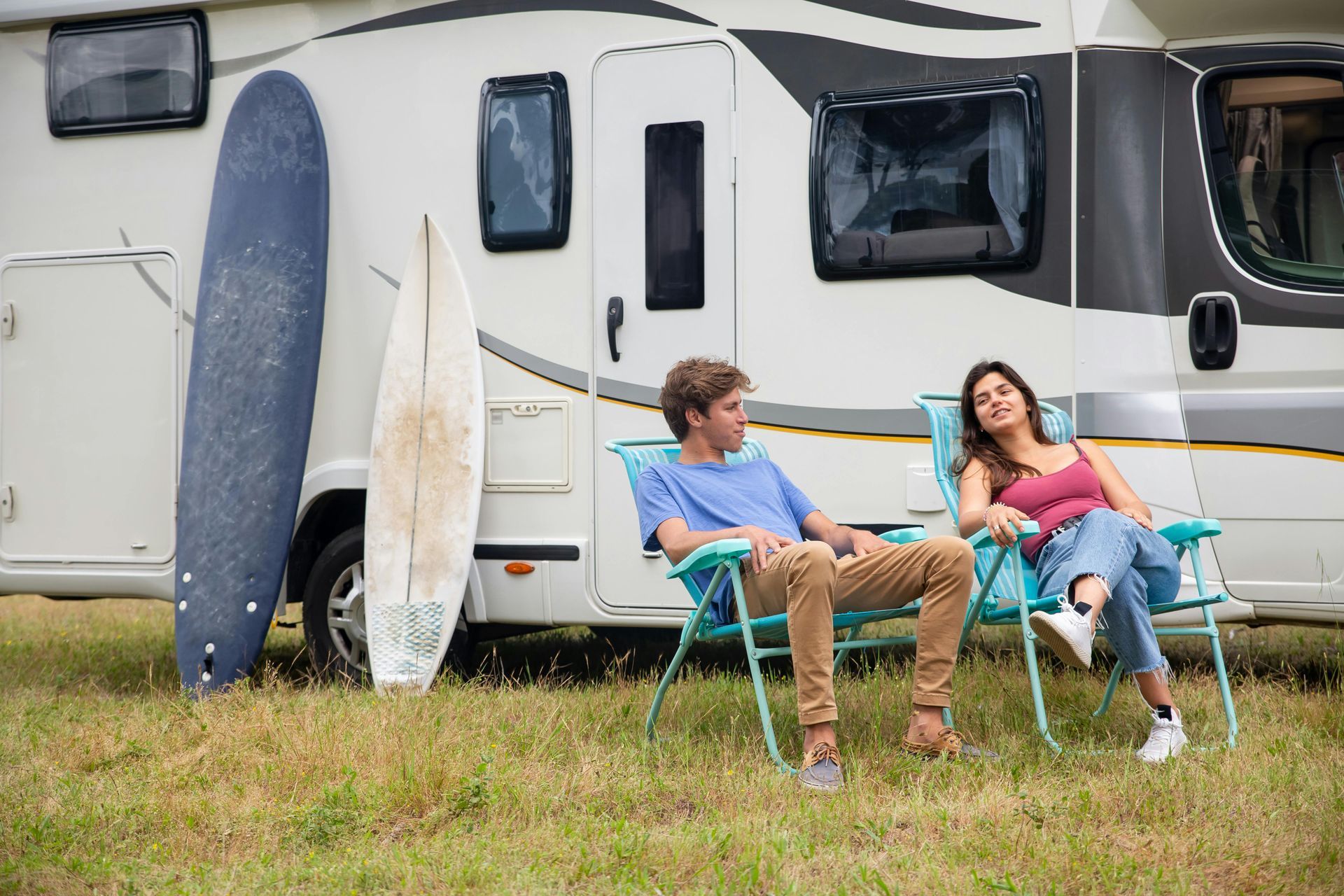 Two people relax in camping chairs outside a parked motorhome with two surfboards leaning against the side.