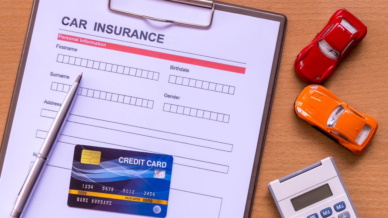A car insurance form with a pen, a credit card, a small calculator, and red and orange toy cars on a wooden surface.