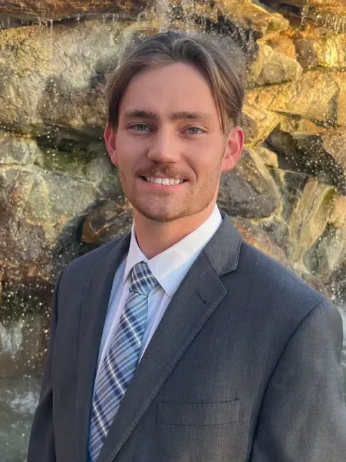 Man in gray suit smiles in front of a rock waterfall. He has blond hair and a blue tie.