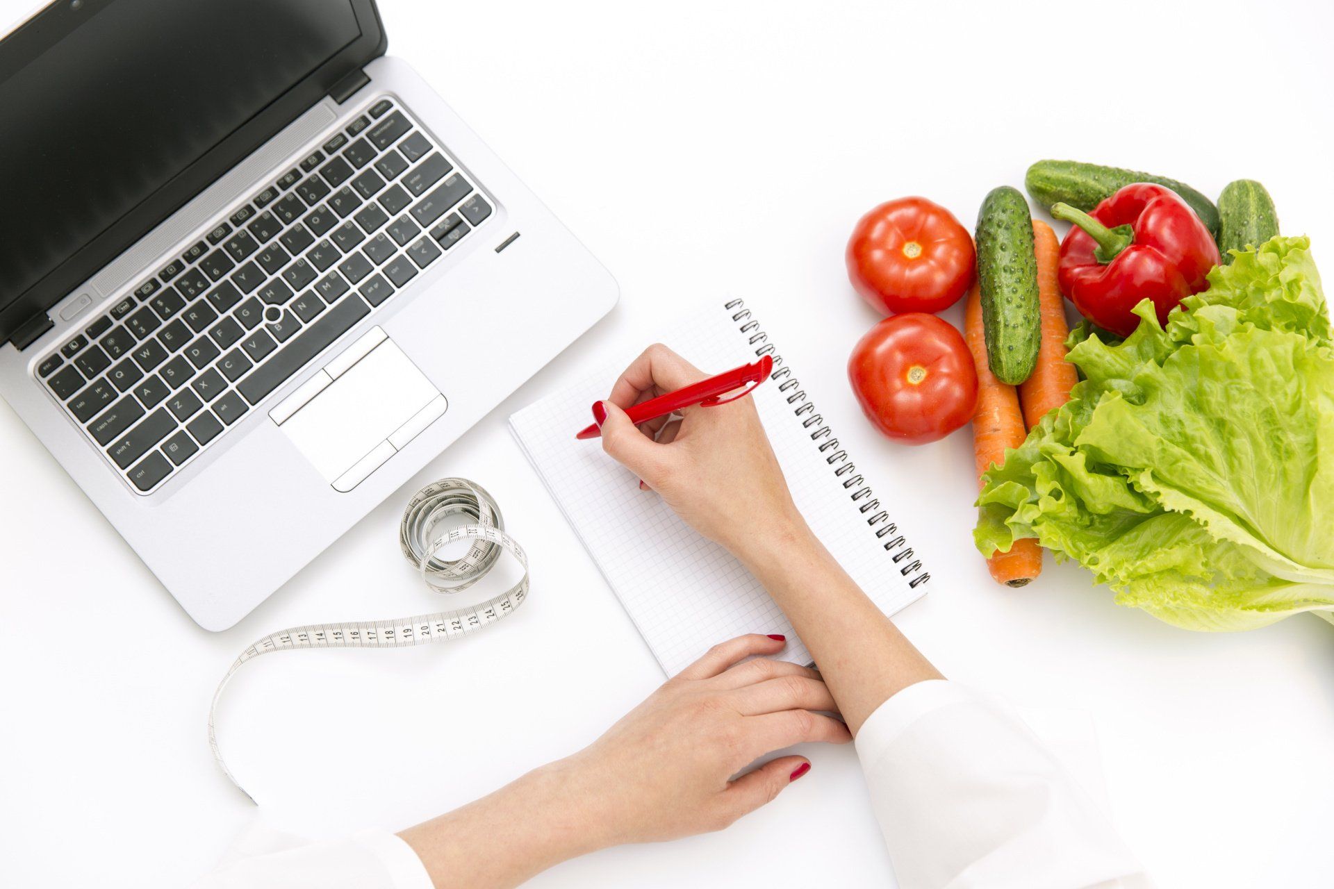 Uma pessoa está escrevendo em um caderno ao lado de um laptop e vegetais.