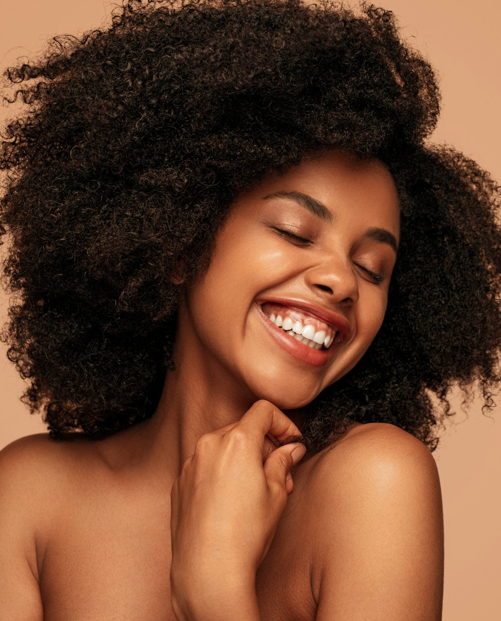 Uma mulher com cabelos cacheados está sorrindo com os olhos fechados.