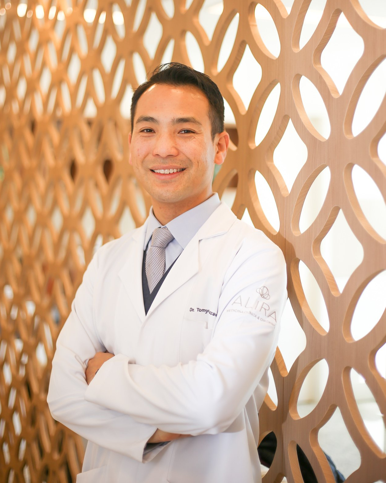 Um homem de jaleco branco e gravata está de pé com os braços cruzados.