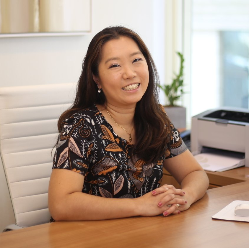 Uma mulher está sentada em uma mesa com as mãos cruzadas e sorrindo