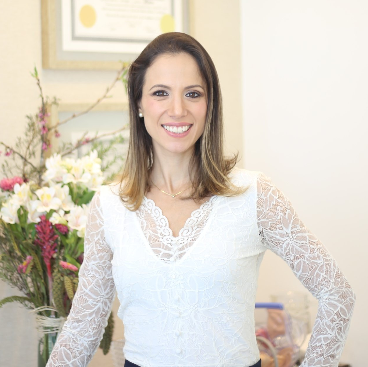 Uma mulher com uma blusa de renda branca está parada em frente a um vaso de flores.
