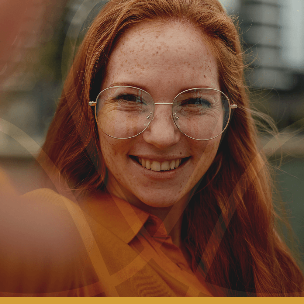 Uma mulher de cabelos ruivos e óculos sorri para a câmera.