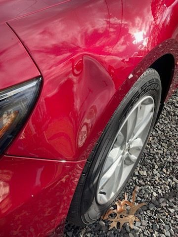 A red car with a damaged fender is parked on gravel.