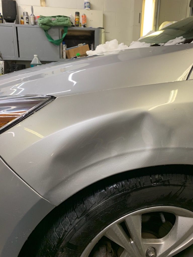 A silver car with a damaged fender is parked in a garage.