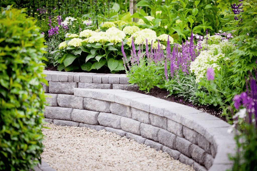 Stone retaining wall with gravel, tiered garden beds, and blooming flowers in various colors.