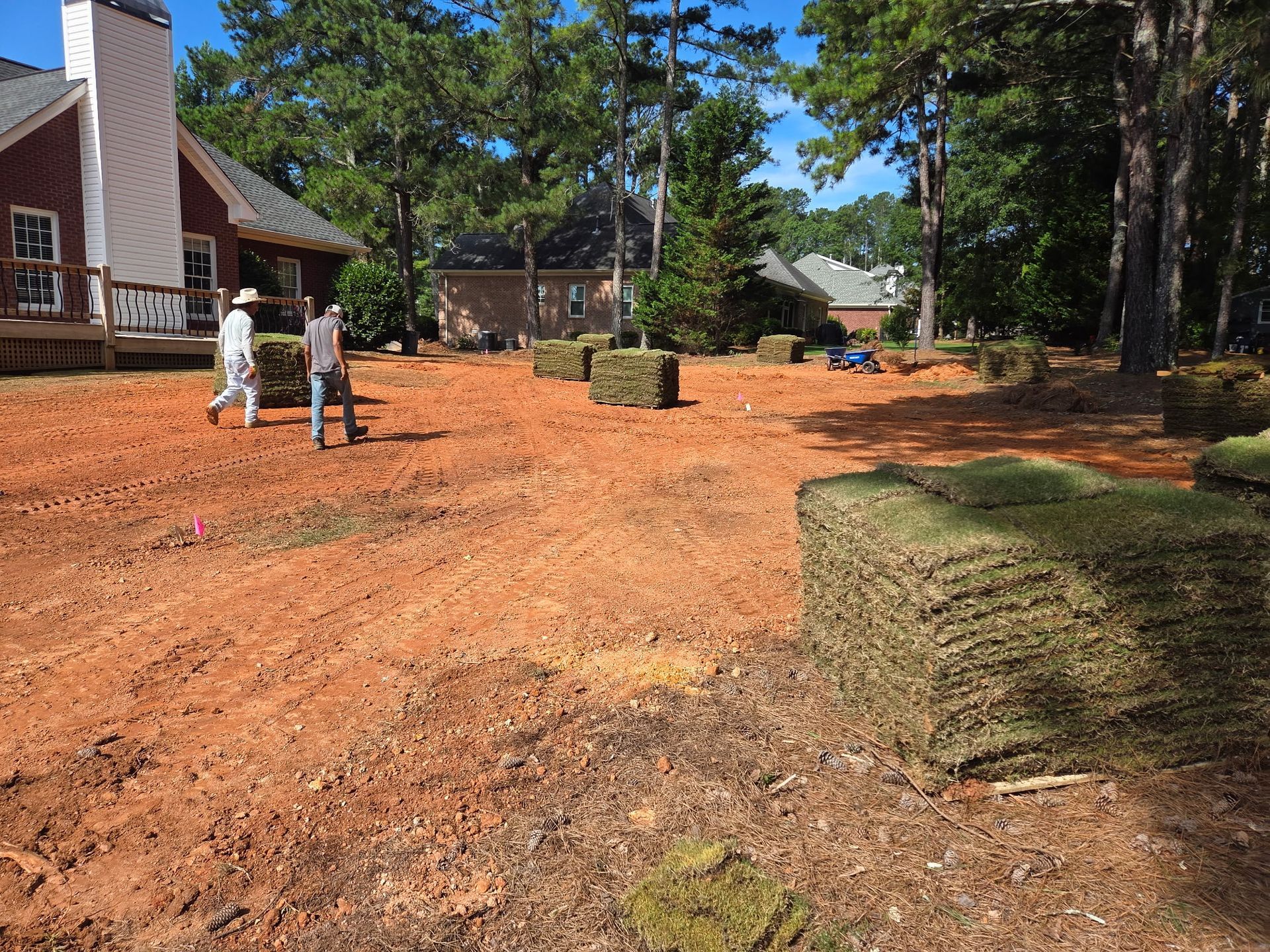 Two people prepare to lay sod in a yard with stacked sod rolls; reddish soil, trees, and houses.
