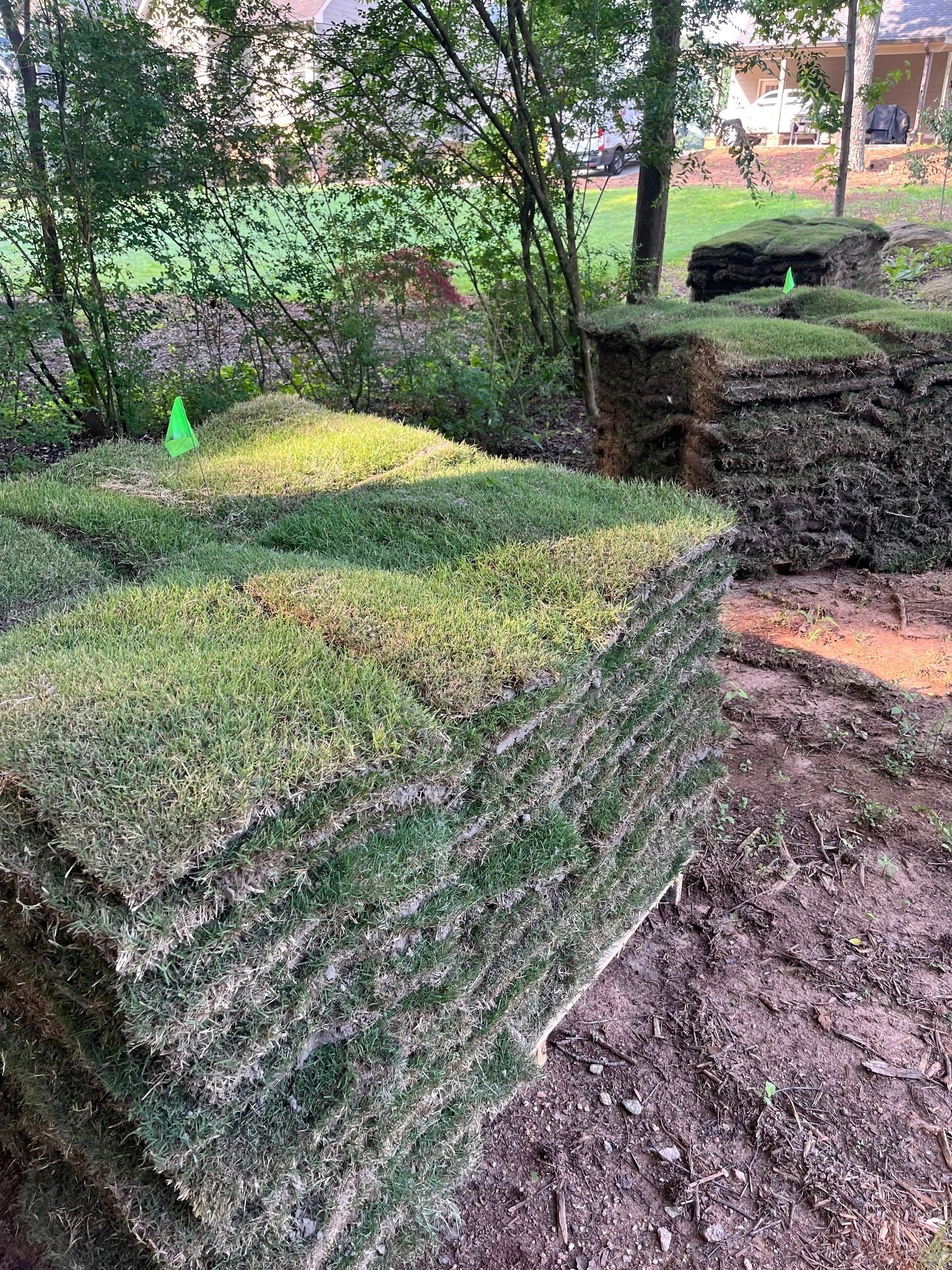 Piles of sod, freshly cut, stacked on bare dirt, with a green marker. Trees and a house in the background.