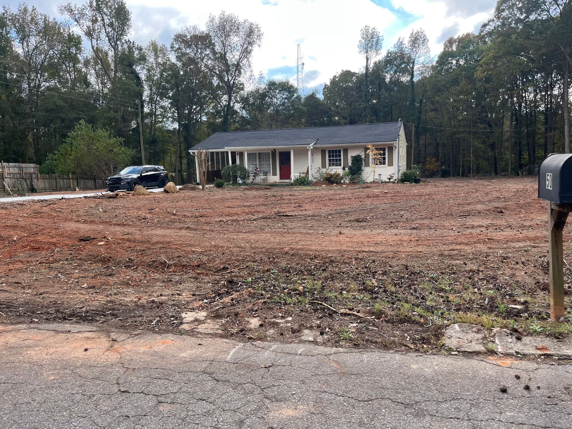 Single-story house with red door, on a cleared lot with a black car visible. Trees in the background.