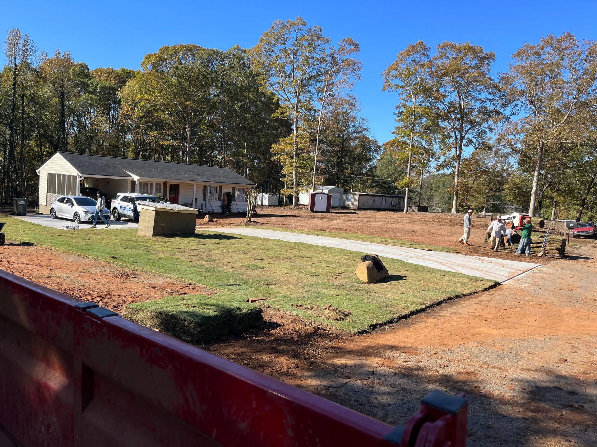 A one-story house with cars in the drive; people work on a lawn area with trees in the background.