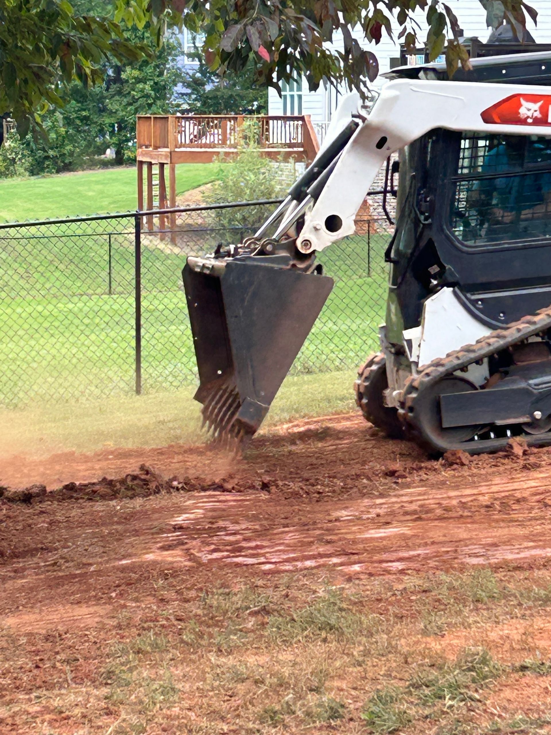 Bobcat skid steer digging a trench in red soil; grassy yard with fence and deck in background.