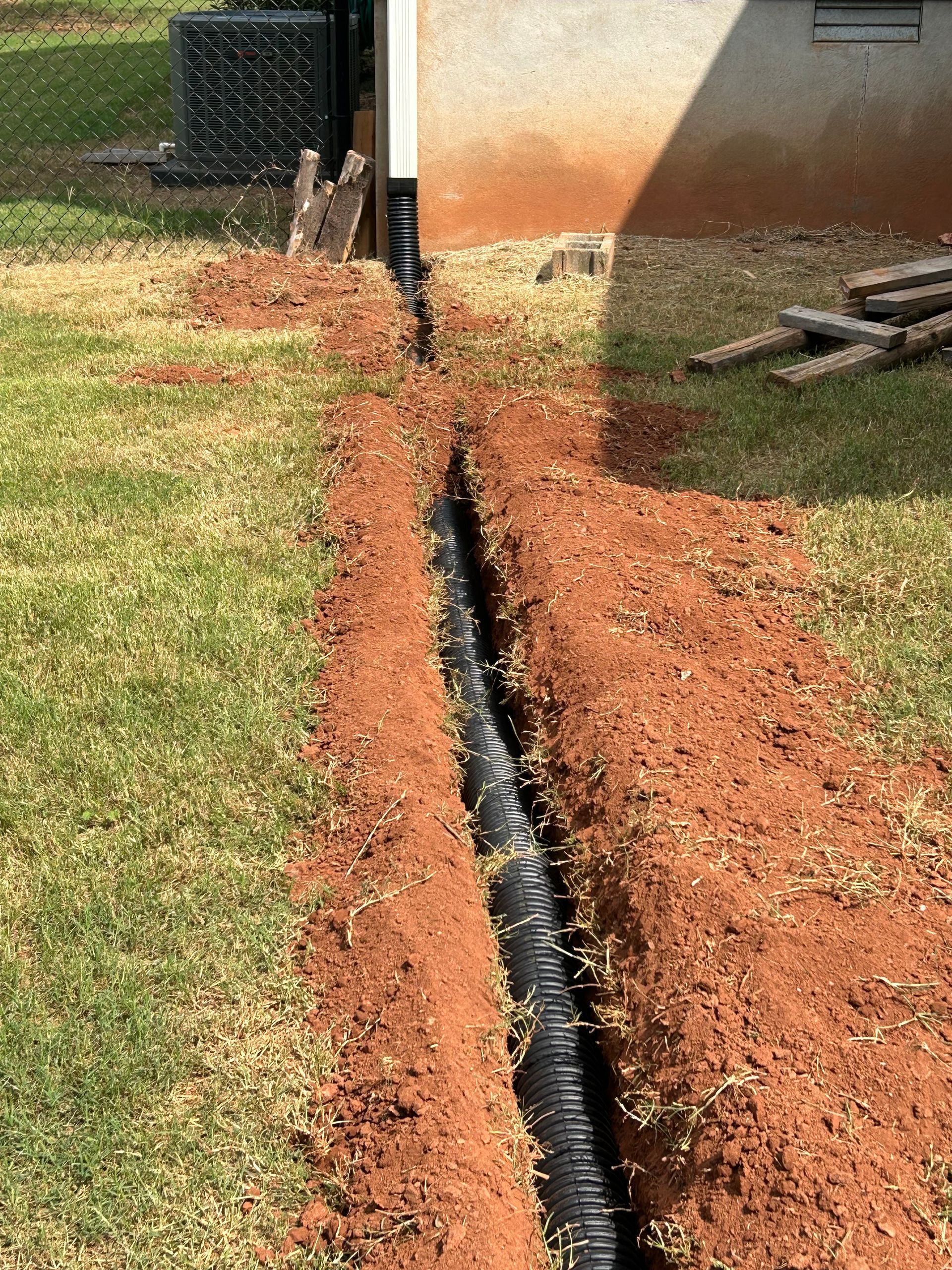 A trench dug in red soil, with black corrugated pipe for drainage, next to a building and lawn.