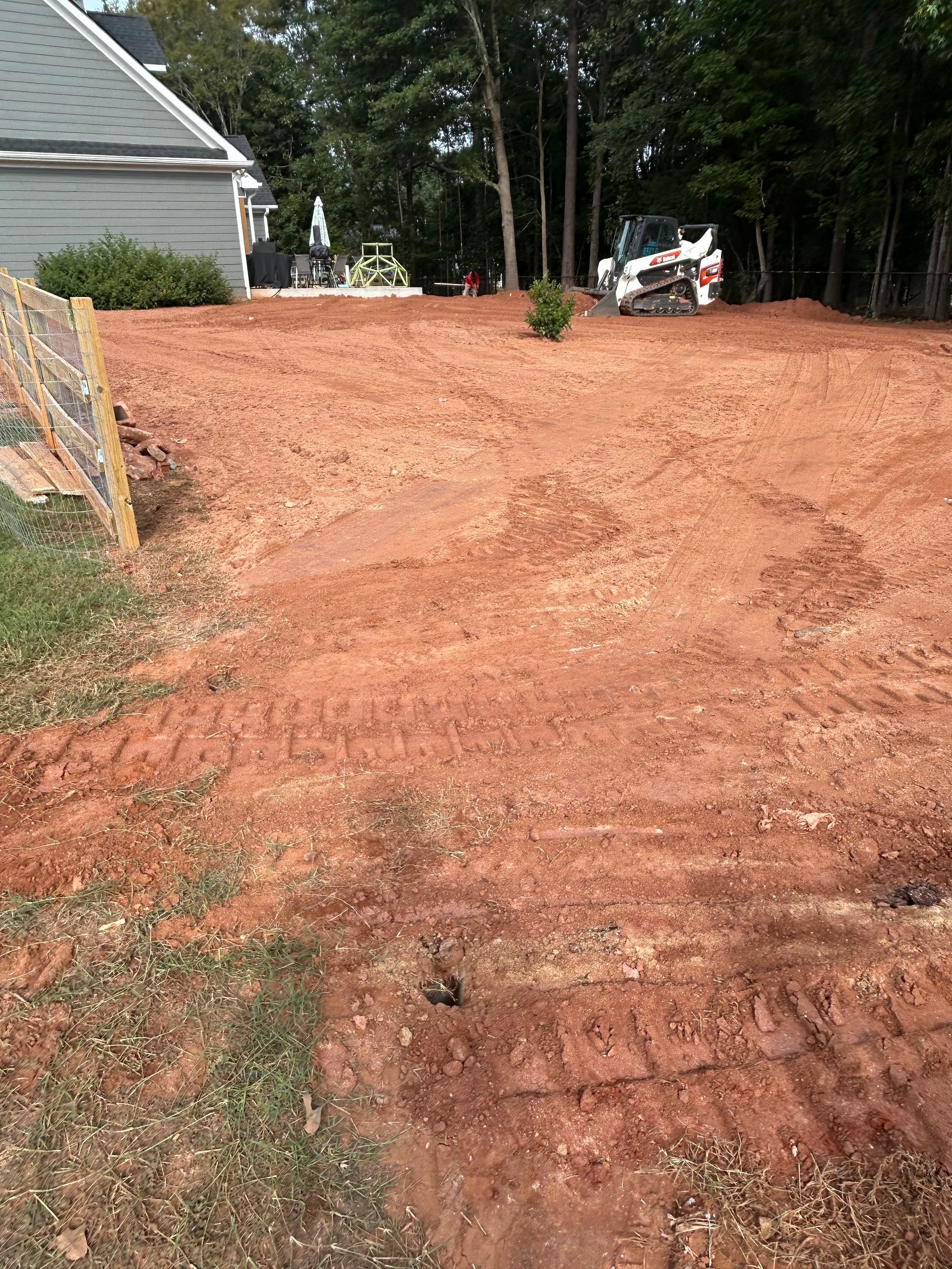 Red dirt yard with a Bobcat, trees, and a house in the background.