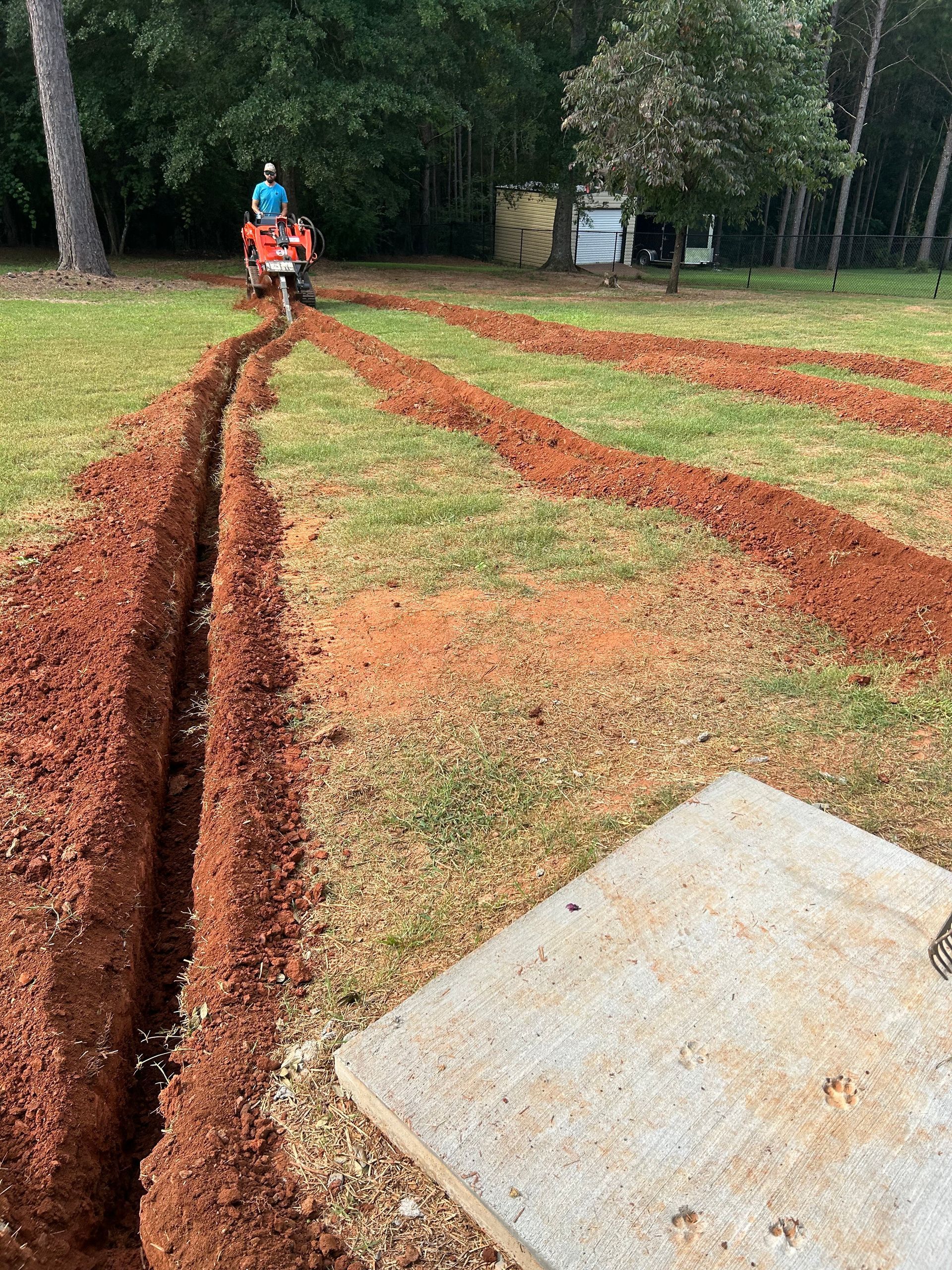 Person on a trencher digging lines in a grassy yard, creating a path of exposed red earth.