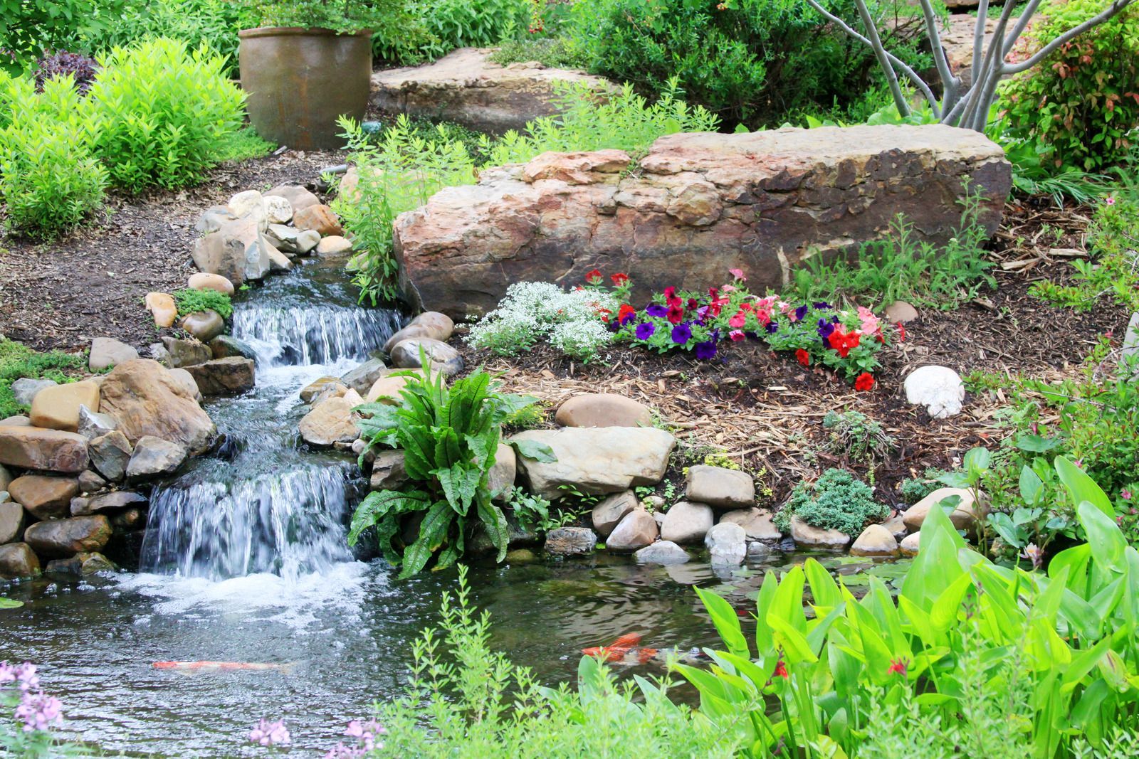 Small waterfall flowing into a pond with plants and colorful flowers, set in a garden.