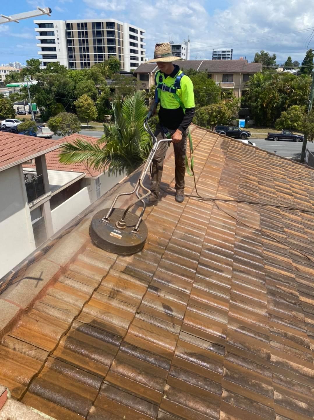 A man cleaning a red tile roof with a pressure washer on a sunny day, city in the background. — SEQ Roof Repaints In Bonogin, QLD