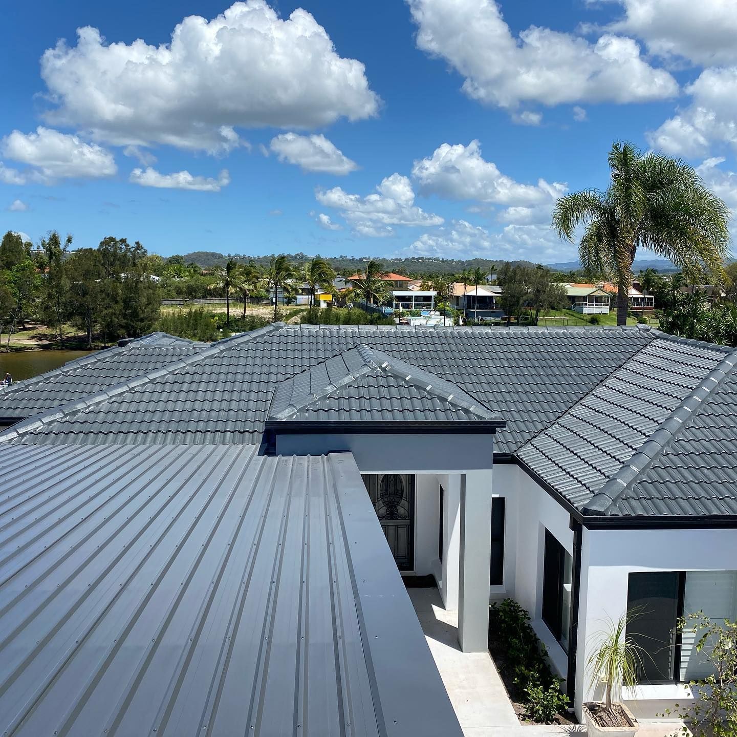 Gray Tile Roof With Blue Sky, White House, and Green Trees — SEQ Roofing & Painting in Carrara, QLD