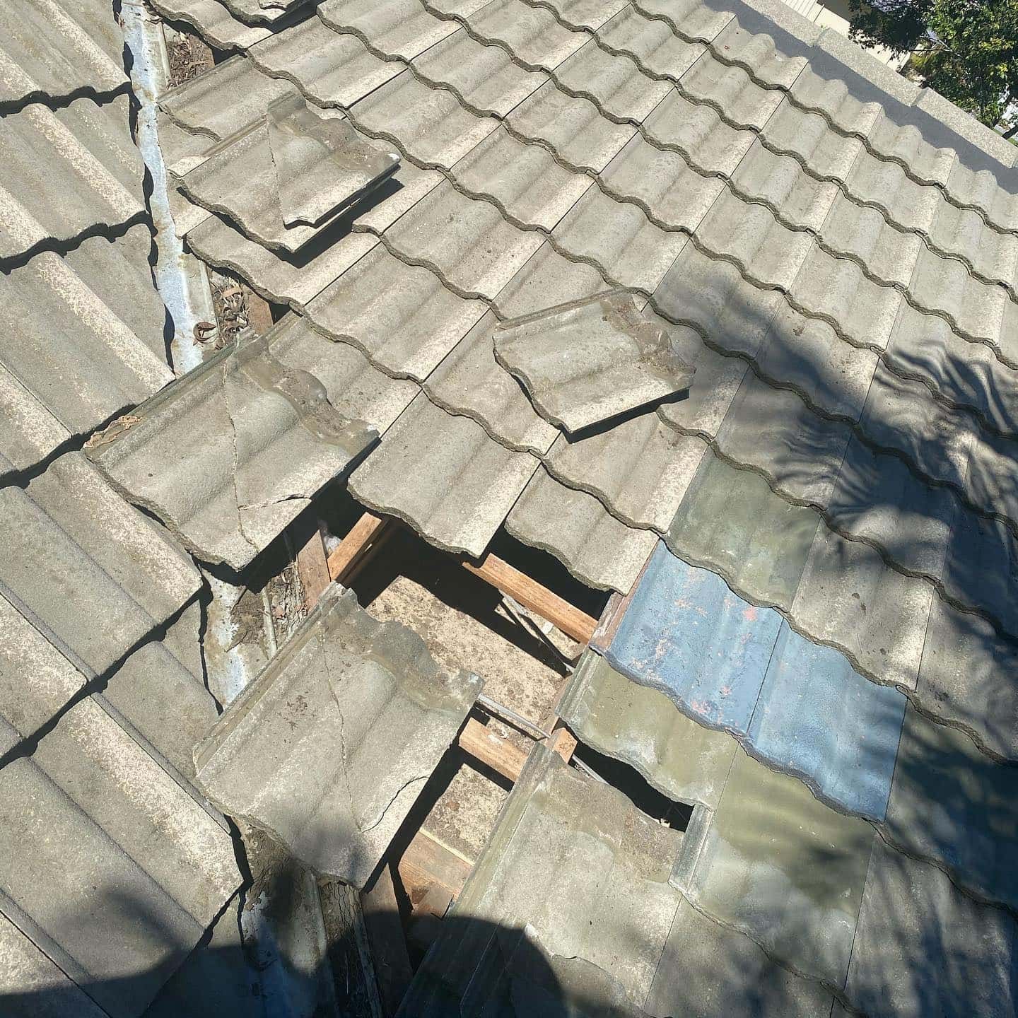 Damaged gray tile roof with missing tiles revealing wooden structure beneath. — SEQ Roof Repaints In Bonogin, QLD
