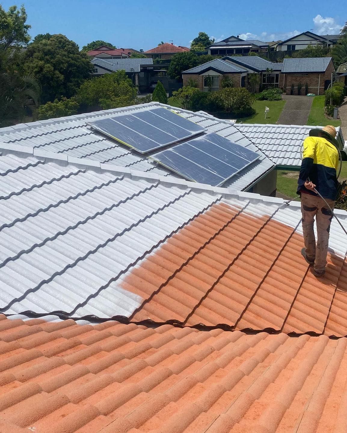 A worker sprays a terracotta roof white near solar panels. Homes are in the background. — SEQ Roof Repaints In Bonogin, QLD