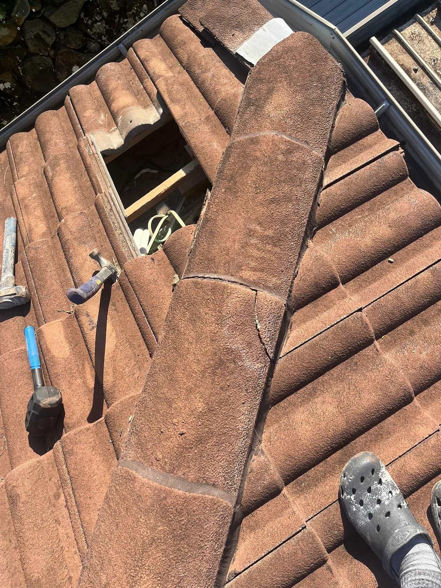 Rooftop with missing tiles exposing interior. Brown tiles, tools, and a foot wearing a Croc. — SEQ Roof Repaints In Nerang, QLD