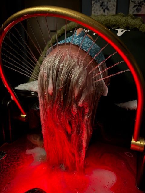 Person's head, hair covered in foam, illuminated by red light. Water runs down a semi-circular structure.