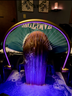 A person lying back receives a head spa treatment, with water flowing from an arched fountain over hair into a basin.