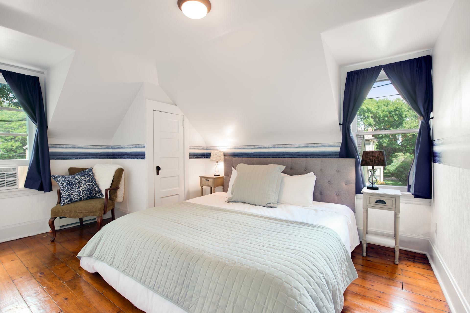 A bedroom with a bed , chair , nightstand and two windows.