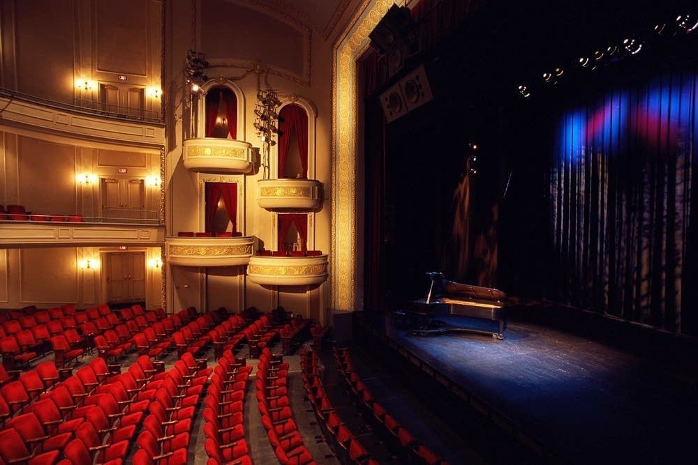 An empty auditorium with red seats and a piano on stage.